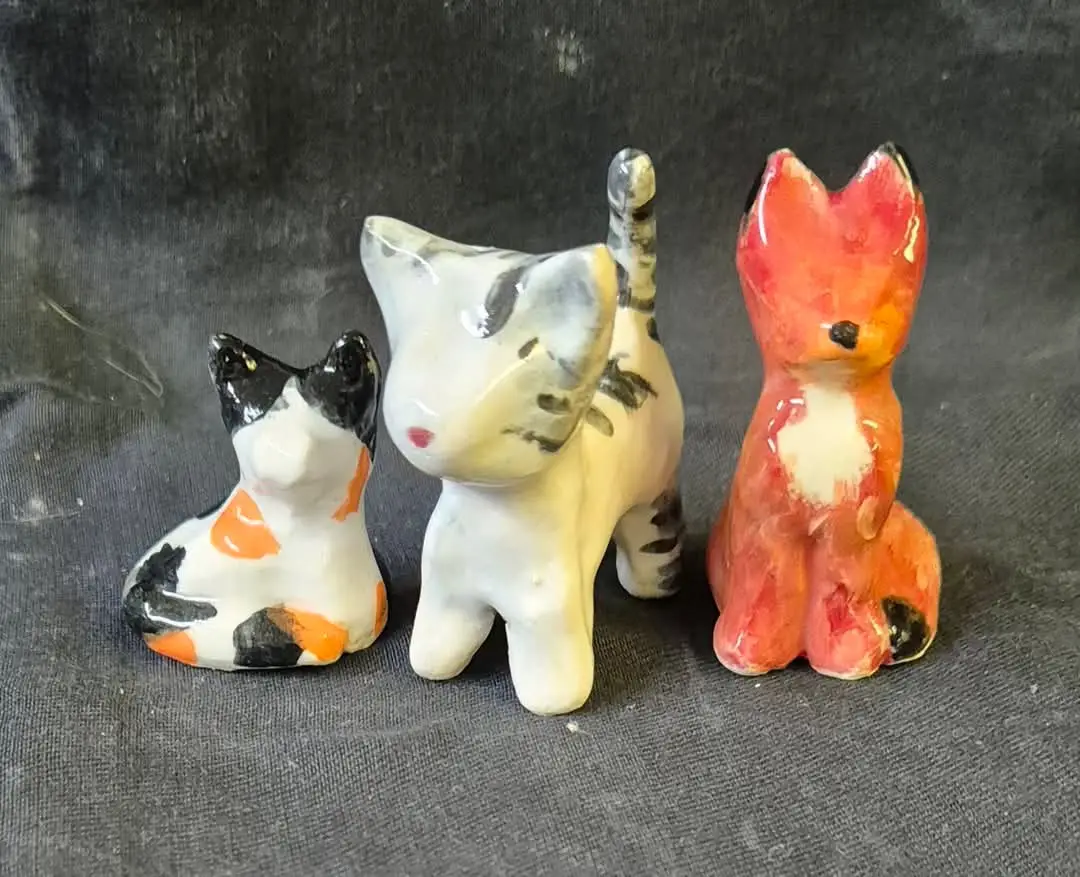 Three cat pottery sculptures made by students at Clay Magic studio, each with unique designs and features.